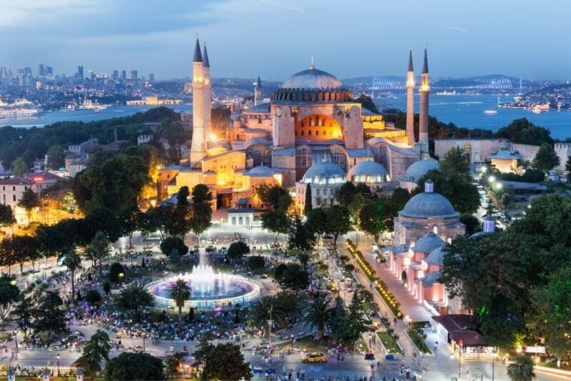 Illuminated Hagia Sophia in Istanbul at dusk, with vibrant city lights, minarets, and a lively park in the foreground.